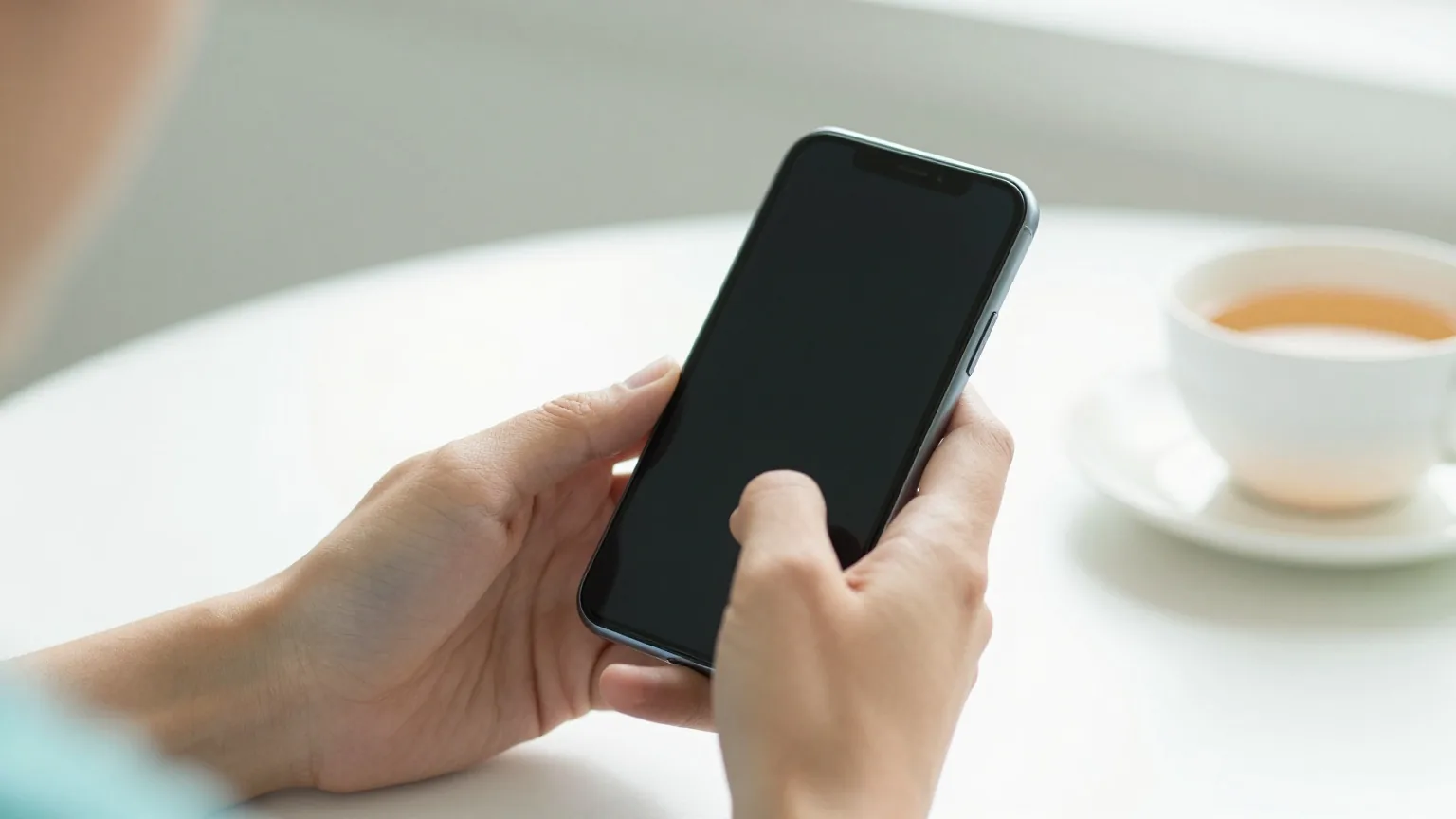 Hände halten ein Smartphone mit abgewandtem Bildschirm an einem hellen Tisch mit einer Tasse Tee