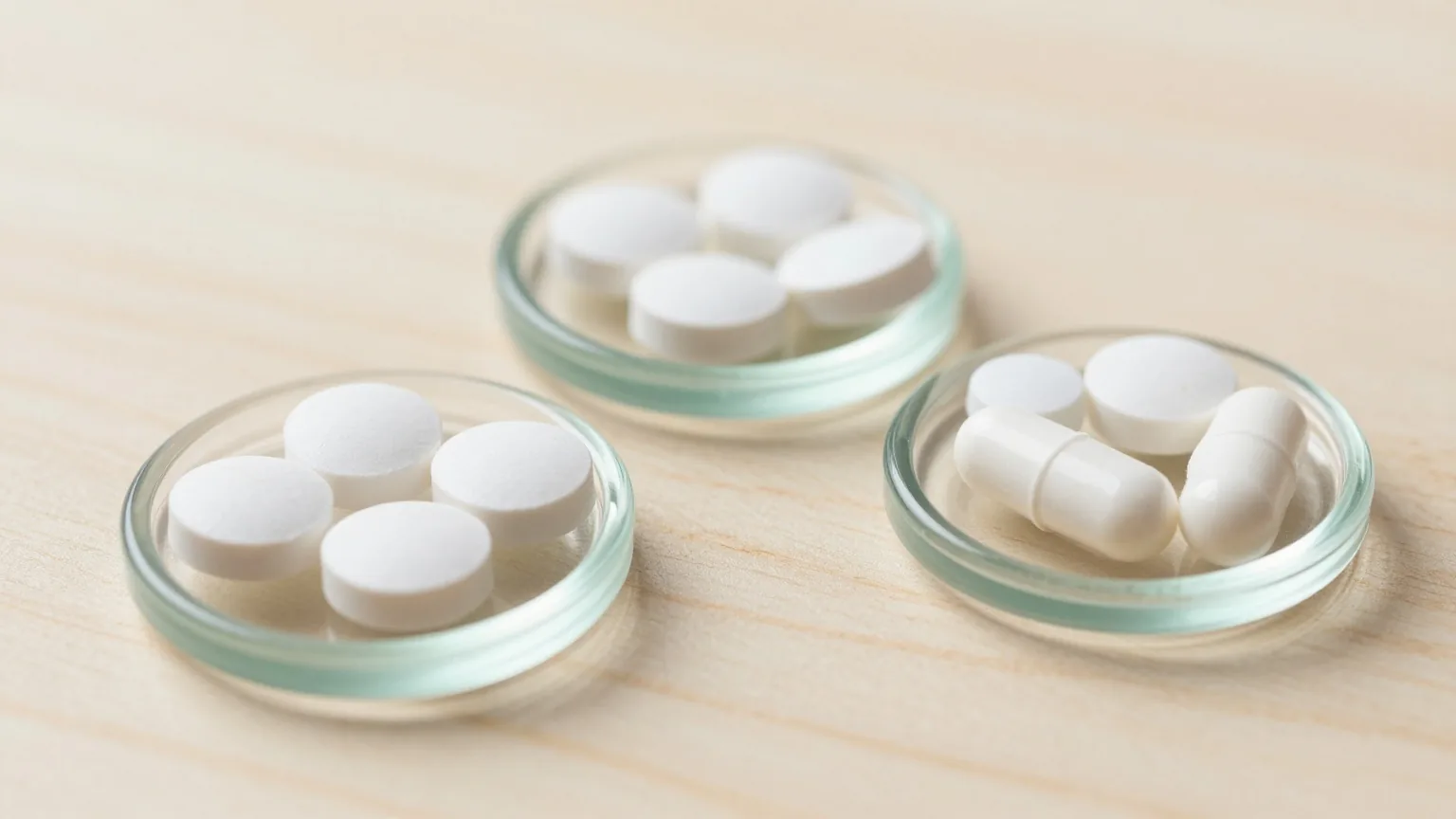 Drei kleine Glasschälchen mit unterschiedlich geformten weißen Tabletten ohne Prägung auf einem hellen Holztisch