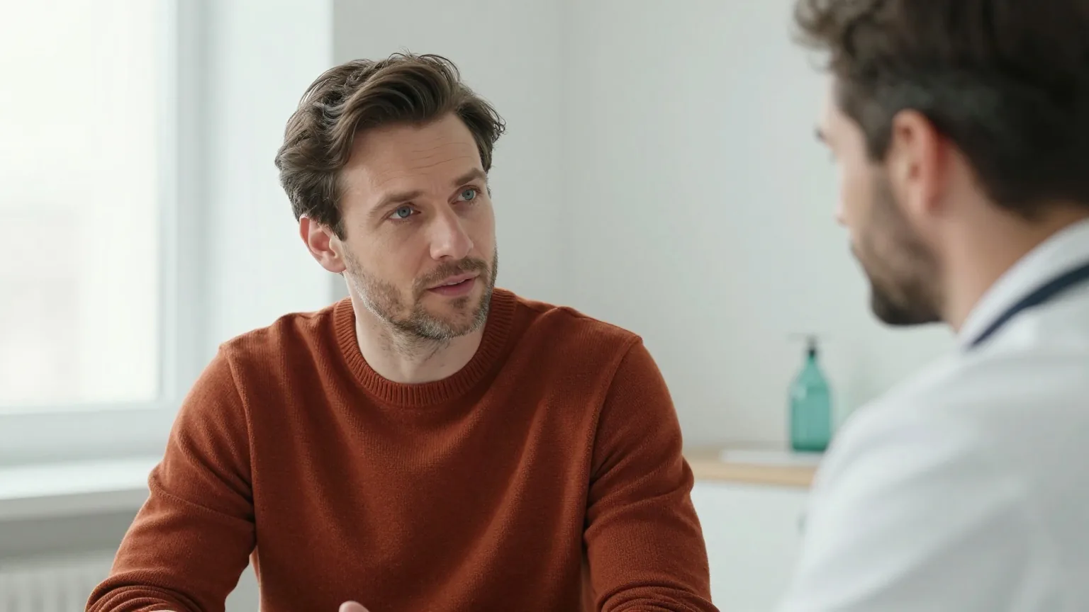 Mann Mitte vierzig mit dunkelbraunem Haar in einem rostroten Pullover sitzt in einem hellen Sprechzimmer einem Arzt mit grauem Bart gegenüber, beide sprechen ruhig miteinander