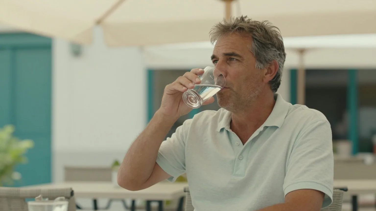 Mann in den Vierzigern sitzt im Schatten einer Markise auf einer Terrasse und trinkt ein Glas Wasser