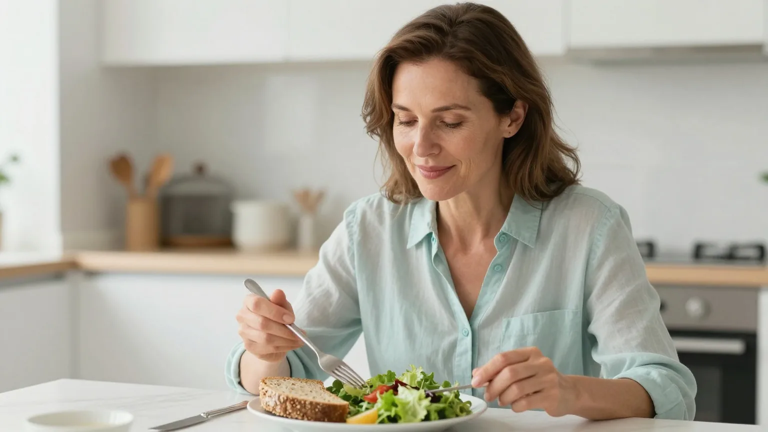 Frau in den Fünfzigern sitzt in einer hellen Küche vor einem Teller mit Vollkornbrot und Salat und beginnt ihr Mittagessen